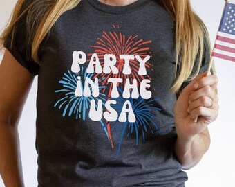 Puede incluir: Una camiseta gris oscuro con el texto "Party in the USA" en blanco con gráficos de fuegos artificiales rojos y azules. La persona sostiene una pequeña bandera estadounidense.