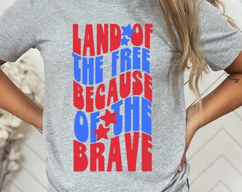 Puede incluir: Una camiseta gris con el texto "Land of the Free Because of the Brave" en letras rojas y azules con estrellas.