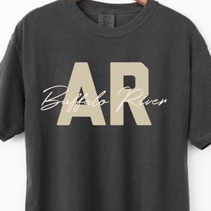 May include: Dark gray t-shirt with the text "AR Buffalo River" in a beige color. The letters "AR" are in a block font, and "Buffalo River" is in a cursive font. The t-shirt is hanging on a wooden hanger.