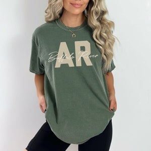 May include: Olive green t-shirt with the letters "AR" in a tan block font and the words "Buffalo River" in a cursive font. The shirt is a short-sleeved, crew neck style.