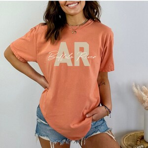 May include: A coral-colored t-shirt with the text "AR Buffalo River" printed on the front. The letters "AR" are in a block font, and "Buffalo River" is in a cursive font. The shirt has short sleeves and a crew neck.