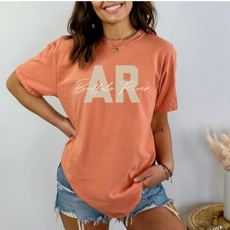 May include: A coral-colored t-shirt with the text "AR Buffalo River" printed on the front. The letters "AR" are in a block font, and "Buffalo River" is in a cursive font. The shirt has short sleeves and a crew neck.