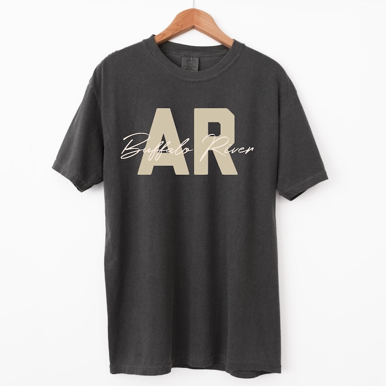 May include: Dark gray t-shirt with the text "AR Buffalo River" in a beige color. The letters "AR" are in a block font, and "Buffalo River" is in a cursive font. The t-shirt is hanging on a wooden hanger.