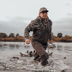 May include: A person wading through water, wearing camouflage hoodie and cap, brown waders, and holding a rope. The person is in a body of water with duck decoys. The sky is overcast.