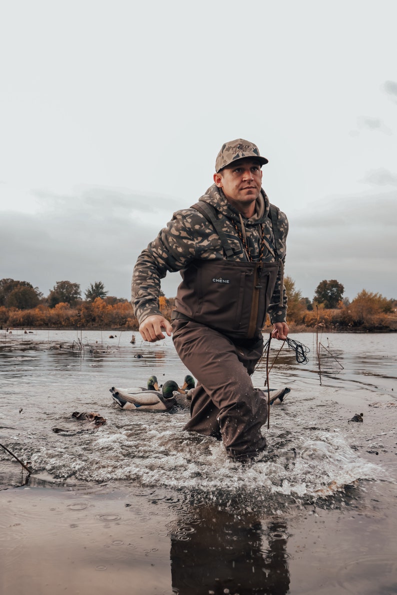 May include: A person wading through water, wearing camouflage hoodie and cap, brown waders, and holding a rope. The person is in a body of water with duck decoys. The sky is overcast.