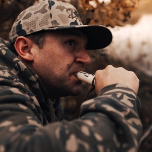 May include: A camouflage baseball cap with a dark brim and a logo is worn by a person. The person is wearing a camouflage hoodie and is holding a white and brown hunting call. The background is blurred with fall foliage.