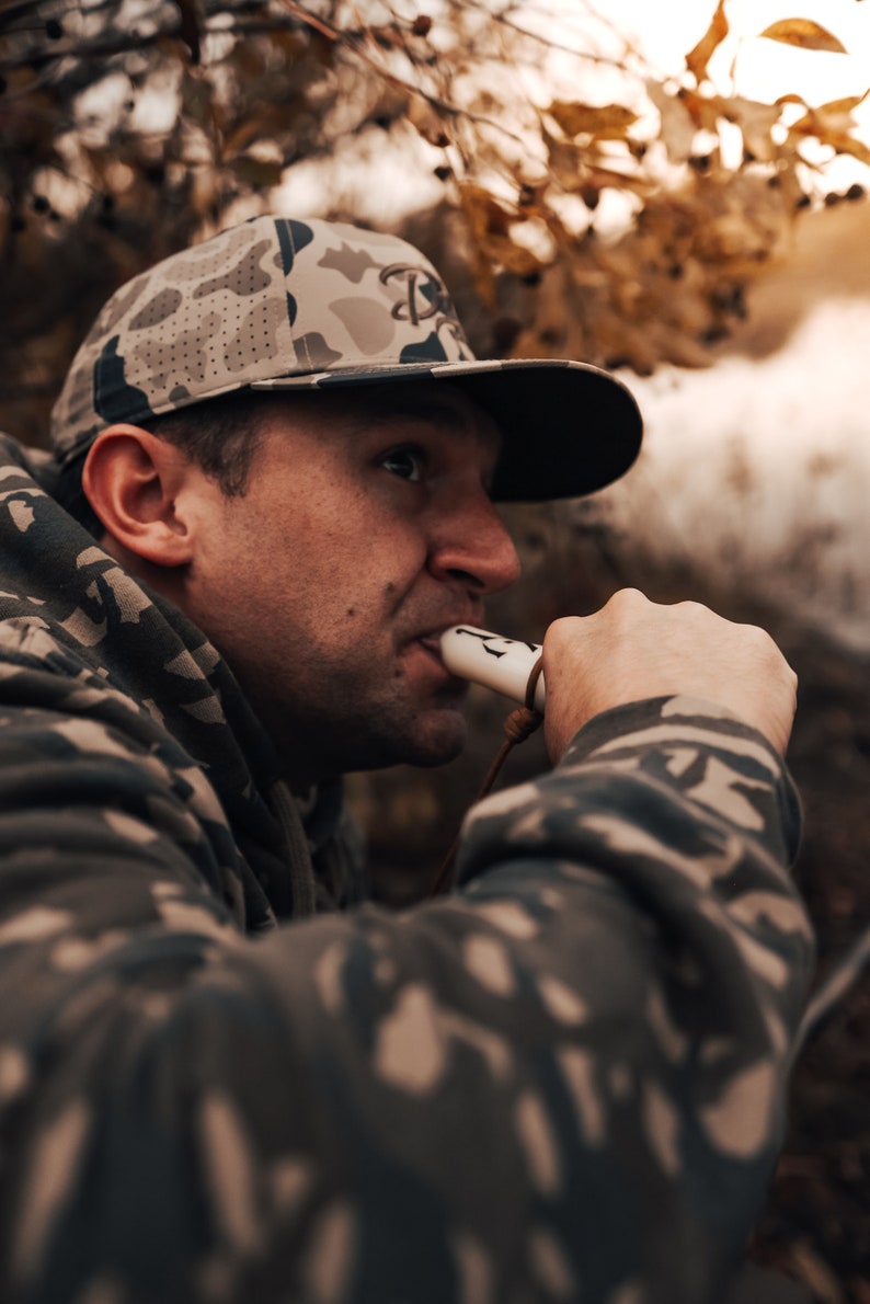 May include: A camouflage baseball cap with a dark brim and a logo is worn by a person. The person is wearing a camouflage hoodie and is holding a white and brown hunting call. The background is blurred with fall foliage.