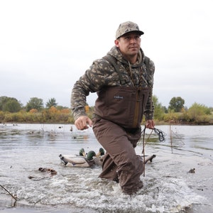 May include: A person wearing camouflage clothing and waders walks through water, holding a rope. The person is in a lake with duck decoys. The clothing includes a camouflage hoodie and a brown wader. The scene suggests duck hunting.