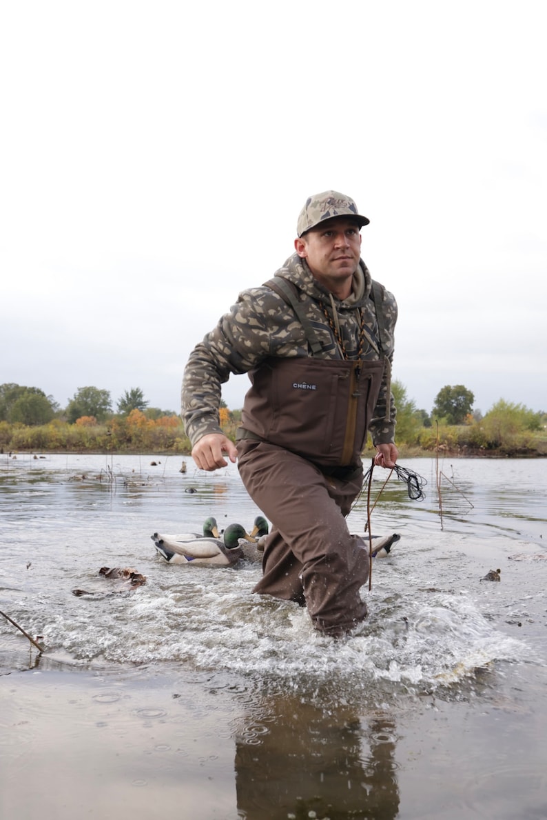 May include: A person wearing camouflage clothing and waders walks through water, holding a rope. The person is in a lake with duck decoys. The clothing includes a camouflage hoodie and a brown wader. The scene suggests duck hunting.