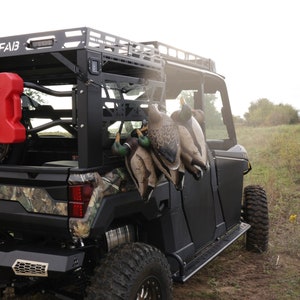 May include: A black utility terrain vehicle (UTV) with a roof rack and large off-road tires. Several duck decoys are hanging on the side. A red fuel container is mounted on the back. The UTV is parked on a dirt path.