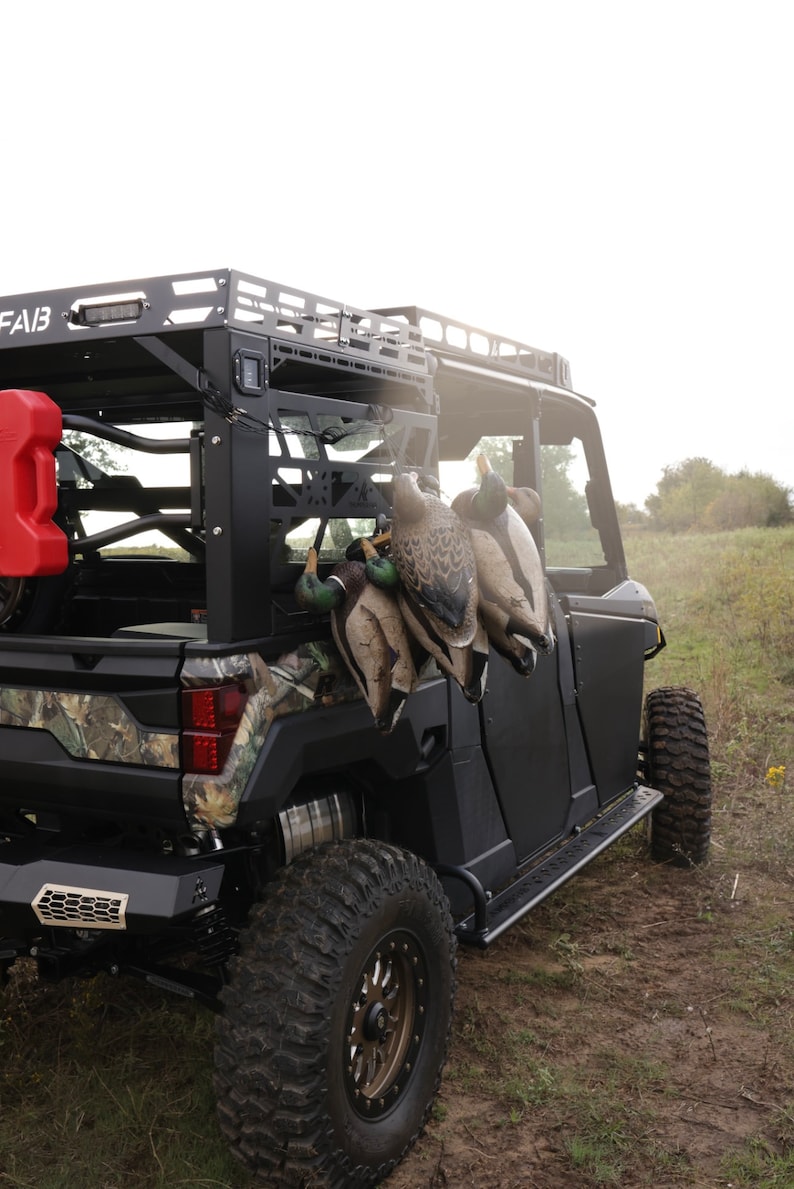 May include: A black utility terrain vehicle (UTV) with a roof rack and large off-road tires. Several duck decoys are hanging on the side. A red fuel container is mounted on the back. The UTV is parked on a dirt path.