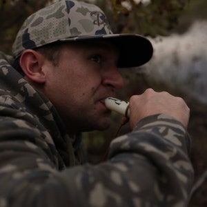 May include: A camouflage baseball cap with a black brim and a logo is worn by a person. The person is wearing a camouflage hoodie and blowing into a white whistle with a brown lanyard. The background is blurred.