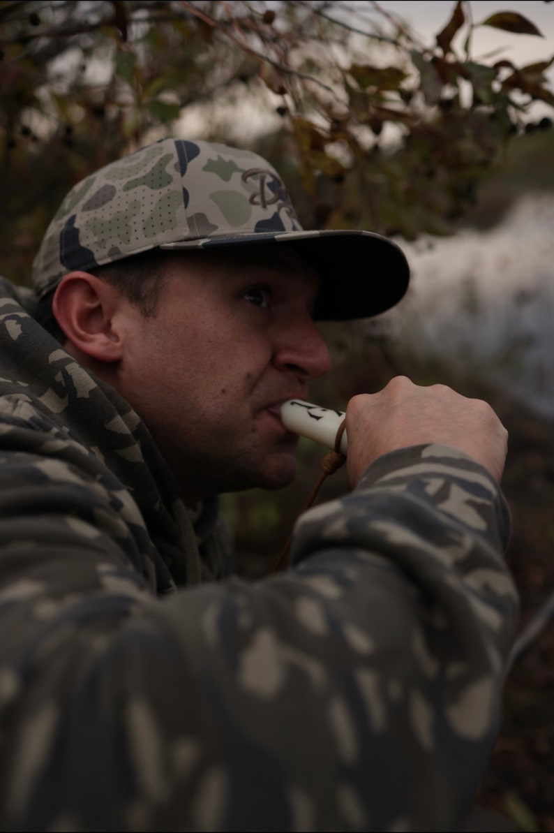 May include: A camouflage baseball cap with a black brim and a logo is worn by a person. The person is wearing a camouflage hoodie and blowing into a white whistle with a brown lanyard. The background is blurred.