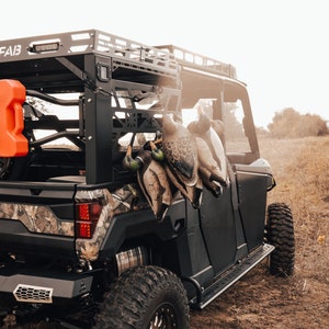 May include: A black off-road vehicle with a camouflage design on the rear. Several hunting decoys are hanging from the vehicle's rack. An orange fuel container is mounted on the side. The vehicle is parked in a field.