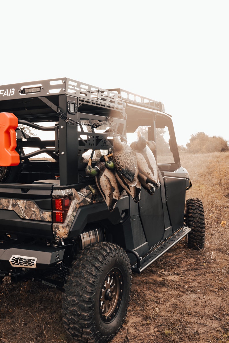 May include: A black off-road vehicle with a camouflage design on the rear. Several hunting decoys are hanging from the vehicle's rack. An orange fuel container is mounted on the side. The vehicle is parked in a field.