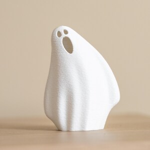 May include: A white ghost figurine with a textured surface. The ghost has two small eye holes and a larger oval mouth opening. The figurine is standing on a wooden surface against a neutral background. The ghost is a Halloween decoration.