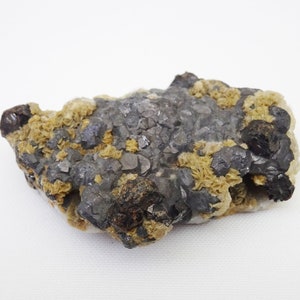 May include: A close-up of a gray and brown mineral specimen with a rough, textured surface. The mineral has a crystalline structure and is surrounded by a white background.