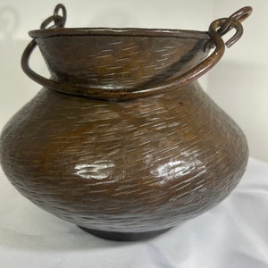 May include: A hammered copper pot with two iron handles. The pot has a rounded body and a wide opening.