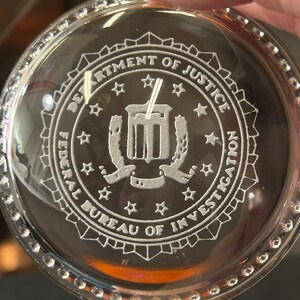 May include: A clear, round glass object with the engraved text "DEPARTMENT OF JUSTICE" and "FEDERAL BUREAU OF INVESTIGATION". The design includes stars and a central emblem. The rim is decorated with small, clear, round embellishments.