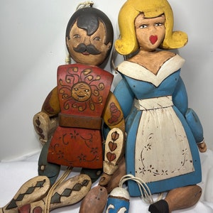 May include: A pair of wooden hand puppets, one with a red and yellow costume and the other with a blue and white dress. The puppets are hanging from strings and have painted faces.