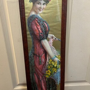 May include: A framed vintage print of a woman in a pink dress with black lace trim. She is wearing a crown of roses and holding a basket of yellow flowers. The background is a gold metallic color.