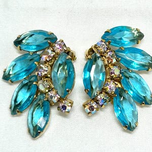 May include: A pair of vintage earrings featuring a cluster design. Each earring has multiple teardrop-shaped, light blue crystals and smaller iridescent crystals set in a gold-tone metal. The earrings are displayed on a white surface.