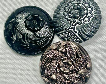 May include: Three decorative buttons with intricate floral and geometric designs. One button is black, another is silver, and the third is a darker silver color. The buttons are round and have detailed patterns.