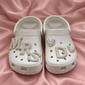 May include: A pair of white clogs featuring decorative charms. The left shoe displays the letters M, R, and S in a pearl and rhinestone finish. The right shoe features a heart-shaped charm, the letter D, and a smaller heart-shaped charm.