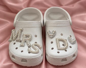 May include: A pair of white clogs featuring decorative charms. The left shoe displays the letters M, R, and S in a pearl and rhinestone finish. The right shoe features a heart-shaped charm, the letter D, and a smaller heart-shaped charm.