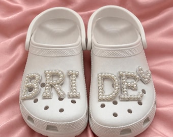 May include: A pair of white slip-on clogs featuring decorative letter charms that spell out the word BRIDE. Each letter is outlined in silver-toned metal and filled with small white faux pearls. A small heart-shaped charm is attached to the letter E.