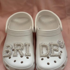 May include: White clogs with the word "BRIDE" spelled out in pearl-like embellishments on one shoe and "DE" with a heart on the other.