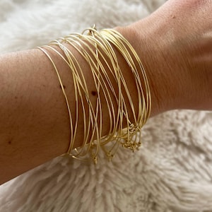 Peut inclure: Un empilement de plusieurs bracelets joncs fins en métal doré poli portés ensemble. Les bracelets sont circulaires, lisses et réfléchissants, créant un aspect superposé autour du poignet avec une épaisseur uniforme et une finition métallique brillante.