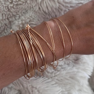 Peut inclure: Un ensemble de multiples bracelets joncs fins en fil de couleur or rose portés ensemble. Les bracelets présentent un design circulaire lisse avec de petits détails noués aux points de fermeture, créant un aspect superposé au poignet.