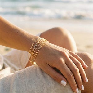 Peut inclure: Un ensemble de cinq bracelets joncs fins en fil doré portés au poignet. Chaque bracelet présente un petit détail de nœud délicat. Les bracelets sont de forme circulaire et ont une finition métallique polie qui reflète la lumière, créant un aspect simple et uniforme.