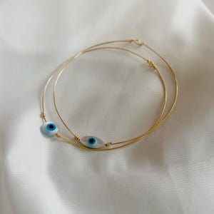 Peut inclure: Deux colliers fins en fil doré, chacun orné d'une perle en verre en forme d'œil protecteur ovale. Les perles sont blanches avec un motif d'iris bleu et noir au centre, maintenues par de petits enroulements de fil doré sur les chaînes.