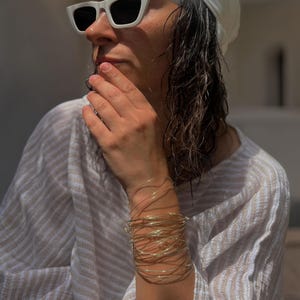 Peut inclure: Un empilement de bracelets fins en fil métallique doré portés au poignet. Les bracelets se composent de multiples brins métalliques délicats et superposés qui créent un aspect circulaire et multicouche autour du bras, reflétant la lumière.