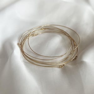 Peut inclure: Un ensemble de bracelets joncs fins en fil métallique doré empilés. Chaque bracelet circulaire présente un petit détail délicat en fil enroulé là où les extrémités se rejoignent, créant un bijou métallique léger et flexible.