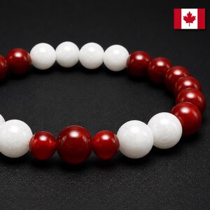 Pode incluir: Uma pulseira de contas com contas redondas vermelhas e brancas alternadas. As contas são esféricas e polidas, dispostas em um padrão repetitivo. A pulseira é enfiada em uma faixa elástica. A bandeira canadense é visível no canto superior direito.