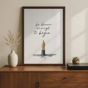 May include: A framed wall art print featuring the black cursive text "be brave enough to begin." centred above a gold-coloured fountain pen nib illustration with a dark grey ink wash accent on a white background, enclosed in a dark brown frame.