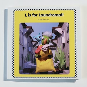 May include: A yellow book cover with a black and white checkered border. The cover features a photo of two puppets in an alleyway. The title of the book is "L is for Laundromat!" by Martha Graft.