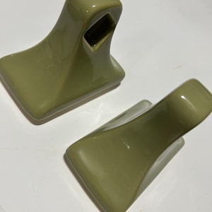 May include: Two olive green ceramic towel holders. Each holder has a rectangular base, a curved body, and a square opening at the top. The holders are glossy and appear to be new.