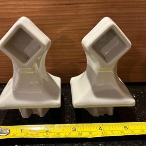 May include: Two white ceramic towel bar holders with a square top and a curved base. The holders are placed side by side on a black surface. A yellow tape measure is placed below the holders, showing measurements in inches.