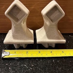 May include: Two speckled, off-white ceramic towel bar holders. Each has a square top and a decorative base. A measuring tape shows the holders are approximately 4 inches wide.