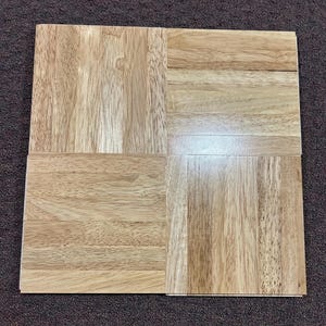 May include: Four square wooden floor tiles arranged in a grid pattern. The tiles are made of light brown wood with visible grain patterns. The wood has a natural finish, showcasing the variations in color and texture.