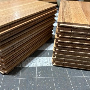 Puede incluir: Dos pilas de tablones de suelo de madera. Los tablones son de color marr&oacute;n claro con un patr&oacute;n de veta de madera. Los tablones est&aacute;n apilados uno encima del otro, creando una forma rectangular.