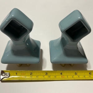 May include: Two light blue ceramic towel holders with a square opening and a base with three small feet. A yellow measuring tape shows the holders are approximately 3 inches wide.