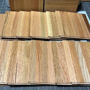 Puede incluir: Una pila de tablones de madera para pisos en varios tonos de marr&oacute;n, con algunos que muestran un patr&oacute;n de veta m&aacute;s claro, casi blanco.