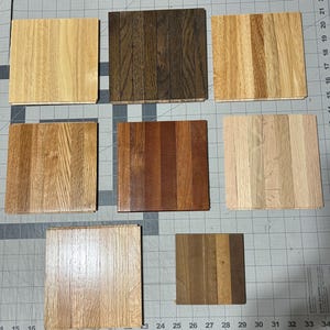 May include: A collection of eight square wood samples showcasing various grain patterns and colors. The samples range from light to dark brown, with visible wood grain details. The squares are arranged on a gray surface with measurement markings.