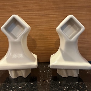 May include: Two off-white ceramic cabinet knobs. Each knob has a square top, a curved body, and a textured base. The knobs are displayed against a wooden background.