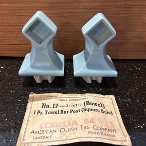 May include: Two light blue ceramic towel bar posts with a square hole design. The posts are from the American Olean Tile Company. The label reads "No. 17 - 2 3/8 x 2 1/2 (Dowel) 1 Pr. Towel Bar Post (Square Hole)".